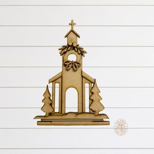 May include: A wooden church cutout with a cross on top, a wreath, and two Christmas trees. The church has an arched doorway and is set on a base. The cutout is made of light brown wood.