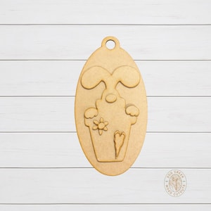 May include: A wooden Easter egg-shaped cutout featuring a bunny and a cupcake design. The bunny has long ears, and the cupcake is decorated with a flower and a heart. The cutout has a hole at the top for hanging.