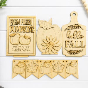 May include: Wooden fall-themed decorations. Includes a sign that reads "Farm Fresh Pumpkins", a pumpkin with a sunflower, a cutting board with "hello FALL", and a banner with pumpkins and leaves.