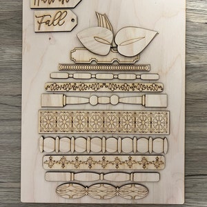 May include: A wooden fall-themed craft kit with laser-cut pieces. Includes a pumpkin shape, decorative borders, and tags that read "Harvest" and "Fall". The pieces are arranged on a wooden base, perfect for DIY projects.