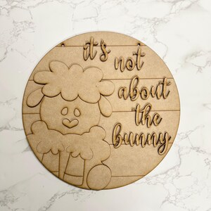 May include: A round wooden sign with a sheep design and the phrase "it's not about the bunny" in cursive. The sign is made of light brown wood and has a horizontal wood grain pattern. Two small holes are at the top for hanging.