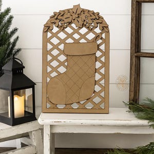 May include: A wooden Christmas stocking decoration with a lattice background and leaf accents. The stocking is tan with a quilted pattern. The decoration is displayed on a white bench next to a black lantern and a small Christmas tree.
