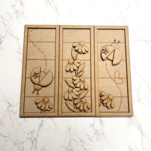 May include: A wooden wall art piece featuring three panels with a nature theme. Each panel has a light brown background with a carved design of a ladybug, flowers, and a dotted line. The ladybugs and flowers are also carved.