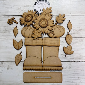 May include: A set of unfinished laser-cut wood pieces featuring a pair of boots with a plaid cuff, sunflowers, pumpkins, an acorn, and various leaves. A circular logo at the top reads Dog Creek Wood Cutouts. Includes a rectangular base stand.