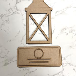 May include: A wooden cutout of a lantern with a rectangular frame and a cross design. The lantern is sitting on top of a rectangular wooden cutout with a circle and two lines.