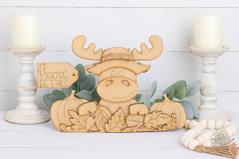 May include: A wooden moose figure wearing a hat, with pumpkins and autumn leaves. The sign says "It Moose Be Fall". Two white candle holders with candles flank the moose.