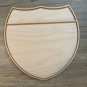 May include: A wooden shield-shaped sign with a light brown wood grain and a dark brown border. The sign has a horizontal line across the middle.