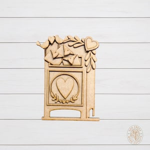 May include: A wooden cutout of a mailbox with a heart-shaped design. The mailbox features a large heart in the center, smaller hearts, birds, and decorative leaves. The cutout is made of light brown wood.