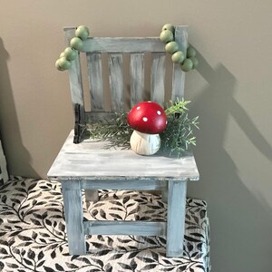 May include: A small, distressed gray wooden chair with a red mushroom decoration and green foliage. The chair has a slatted back and is adorned with green bead garland. The chair is placed on a patterned fabric.