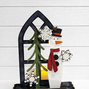 May include: A wooden snowman decoration with a black top hat, red scarf, and carrot nose. The snowman stands in front of a dark blue window frame with a green tree and snowflakes. A small candle is inside the window.