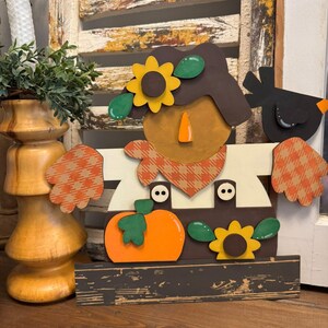 May include: A wooden fall-themed decoration featuring a scarecrow design. The scarecrow has a brown hat, a round face, and a plaid scarf. It is adorned with a pumpkin, sunflowers, and a black bird. The base is a distressed dark brown.