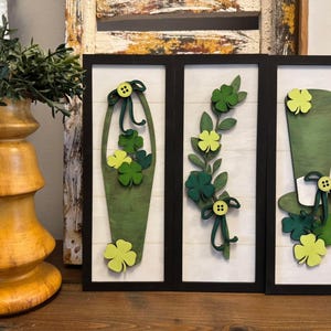 May include: Three framed wood wall decor pieces with a St. Patrick's Day theme. The pieces feature green shamrocks, a green top hat, and a green basket of shamrocks.