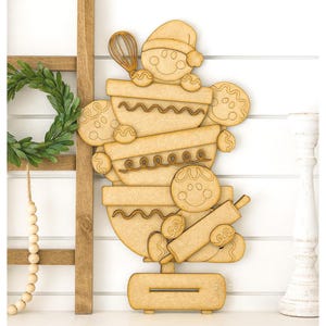 May include: Wooden gingerbread man-themed decoration. The decoration features a stack of mixing bowls, gingerbread men, and cookies. A whisk and rolling pin are also included in the design. The gingerbread man on top is wearing a Santa hat. The decoration is made of light brown wood.