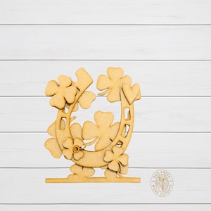 May include: A wooden horseshoe cutout decorated with four-leaf clovers. The horseshoe is surrounded by several clovers and is mounted on a base. The cutout is made of light-colored wood and is suitable for St. Patrick's Day decorations.
