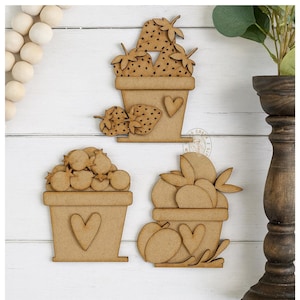 May include: Three wooden cutouts of fruit-filled pots. One pot has strawberries, another has small round fruits, and the third has peaches. Each pot has a heart shape cut out on the front. The cutouts are light brown and sit on a white wooden surface.