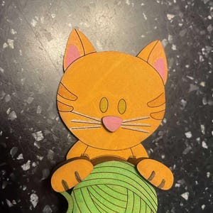 May include: A wooden cutout of an orange cat holding a green ball of yarn. The cat has pink inner ears, a pink nose, and white whiskers. The cat's eyes are oval-shaped and yellow.