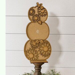 May include: A wooden, three-tiered pumpkin-shaped decoration. The top and bottom sections feature sunflower cutouts. The decoration is mounted on a dark brown, decorative stand. The background is a white, horizontal-slat wall.