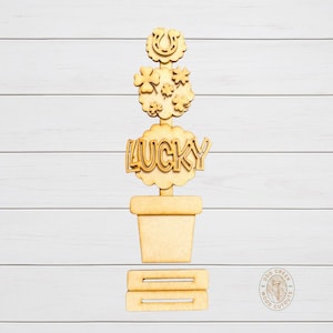 May include: A wooden St. Patrick's Day decoration featuring a potted plant design. The plant has a horseshoe, shamrocks, and the word "LUCKY" cut out. The base includes two horizontal bars. The design is made of light-colored wood.