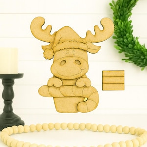 May include: A wooden moose wearing a Santa hat and holding a candy cane. The moose is light brown and has a smiling face. A small wooden block is next to the moose. A candle holder and a beaded garland are in the background.