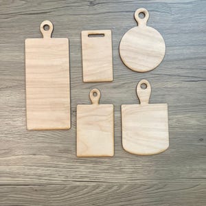 May include: Five unfinished wooden cutting boards in various shapes. The boards include a long rectangle, a small rectangle, a circle, a square, and a rounded rectangle. Each board has a handle. The wood grain is visible.