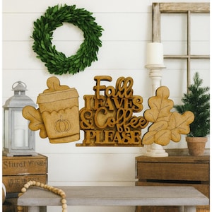 May include: Wooden fall-themed decorations, including a coffee cup with a pumpkin, oak leaves, and a sign that reads "Fall Leaves & Coffee PLEASE." A green wreath hangs above the decorations. The items are displayed on a white wall.