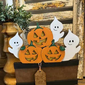 May include: A wooden Halloween decoration featuring three orange jack-o'-lanterns and two white ghost cutouts. The pumpkins have carved faces and green stems. The decoration sits in a brown box with the text "Just Here For The Boos" on a tag.