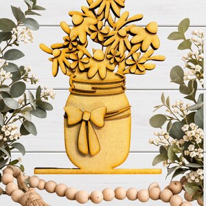 May include: Wooden cutout of a mason jar filled with flowers, with a bow on the front. The flowers and jar are a light yellow color. The cutout is on a white wooden background, with greenery and a beaded garland.