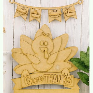 May include: Wooden Thanksgiving decoration featuring a turkey with outstretched wings and a banner that reads "Give Thanks." Above the turkey is a banner with pie-shaped pennants. The decoration is made of light brown wood and is suitable for seasonal decor.