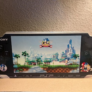 Puede incluir: Una consola de juegos portátil Sony PlayStation Portable (PSP) negra y plateada con una pantalla que muestra un videojuego con Sonic the Hedgehog. El juego se llama "Sonic Advance 3".