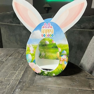 May include: An Easter-themed decorative display featuring a large egg shape with a cutout, bunny ears, and a colorful landscape scene. The words "Happy Easter" are printed on the front. The display is made of cardboard.