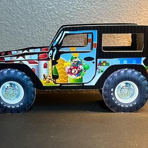 May include: Black toy jeep with a Super Mario Bros. themed design. The jeep has a black body with a blue and white design on the side. The design features Mario, Luigi, Peach, and Yoshi. The jeep has black wheels with silver rims and the words "Goodyear" on the tires.