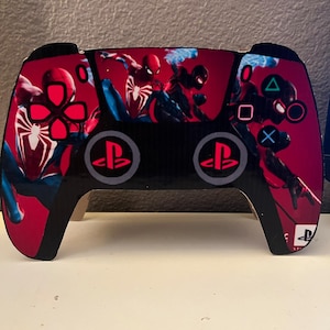 May include: A black PlayStation 5 controller with a red and black Spider-Man themed skin. The controller features the PlayStation logo and the Spider-Man character.