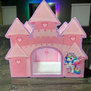 Può includere: Un castello rosa di cartone con ritagli a forma di cuore e una grafica di unicorno. Il castello ha più torrette e un arco centrale. L'unicorno ha criniera, coda e ali arcobaleno. Il castello è su una superficie di legno scuro.