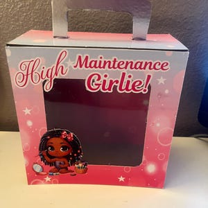 May include: A pink and blue cardboard box with a silver handle. The box has a window that shows a cartoon illustration of a Black girl with braids, wearing a pink bikini top and applying makeup. The text on the box reads "High Maintenance Girlie!"