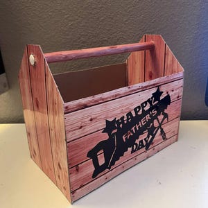 Könnte beinhalten: Eine braune Pappwerkzeugbox mit einem Holzgriff. Die Werkzeugbox hat ein Holzmaserungsmuster und sagt "Happy Father's Day" mit einer Silhouette von Werkzeugen.