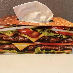 Może przedstawiać: Prostokątne pudełko na chusteczki z nadrukiem podwójnego cheeseburgera, w tym bułki sezamowej, sałaty, pomidora, sera i kotleta. Białe chusteczki wystają z górnego otworu.