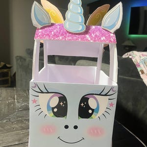 Puede incluir: Una caja de almacenamiento con temática de unicornio. La caja presenta una cara azul claro con ojos grandes y coloridos, pestañas y una boca sonriente. Tiene una parte superior rosa brillante con un cuerno y orejas azules.