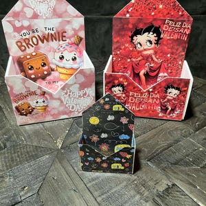 Peut inclure: Trois boîtes décoratives pour la Saint-Valentin. Deux boîtes présentent des motifs roses et rouges avec des personnages de dessins animés et le texte "Happy Valentine's Day" et "You're the Brownie". La troisième boîte a un fond noir avec des illustrations colorées.