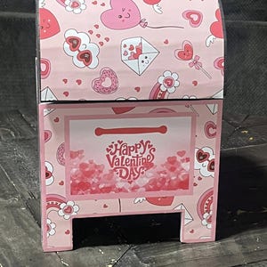 Könnte beinhalten: Ein rosa Valentinstagsbriefkasten mit gewölbter Oberseite. Der Briefkasten ist mit Herzen, Regenbogen und anderen Grafiken zum Valentinstag verziert. Auf der Vorderseite des Briefkastens steht der Text "Happy Valentine's Day".