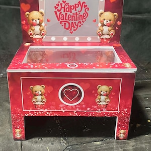 Könnte beinhalten: Eine rote Geschenkbox zum Valentinstag mit einem transparenten Deckel und einem dekorativen Design. Die Box zeigt Teddybär-Illustrationen und den Text "Happy Valentine's Day" in einem herzförmigen Design. Die Box hat ein herzförmiges Logo.
