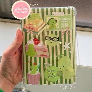 May include: Clear phone case with a green and white striped background, decorated with cartoon frog and cake stickers. The case has the text "MAKE ART WHY NOT" on a pink circle.