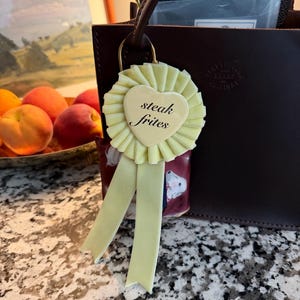 May include: A light yellow rosette-shaped charm with a heart-shaped center that reads "steak frites" in black text. The charm has two long, light yellow ribbons and is attached to a dark brown leather bag.