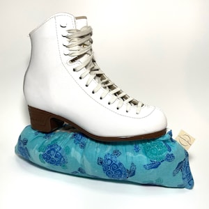 May include: White figure skates with white laces on top of a blue and green fabric skate guard with a sea turtle pattern. The skate guard has a white tag with the text "Charming Threads" on it.