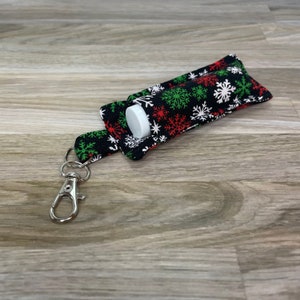 May include: A black fabric keychain with a white, red, and green snowflake pattern. The keychain has a silver clip and a small pocket for a lip balm or other small item.