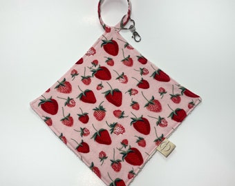 May include: A square fabric cloth with a light pink background featuring a repeating pattern of red strawberries and green stems. The cloth has a matching fabric loop at the top attached to a silver metal lobster clasp and a small beige brand tag.