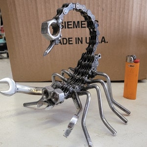 May include: A metal sculpture of a scorpion made from recycled materials, including a wrench and chain. The scorpion is posed with its tail raised and its claws extended. The sculpture is painted in a silver color.