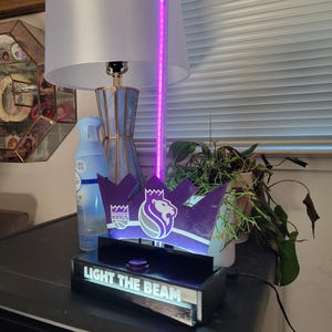 May include: A purple and black decorative light fixture featuring the Sacramento Kings logo and the text "LIGHT THE BEAM". The fixture has a crown design and a purple LED light.