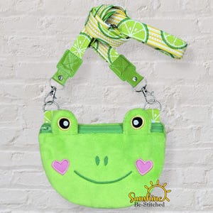 May include: A lime green frog-shaped crossbody bag with a zip closure and a lime slice patterned strap. The frog has pink heart cheeks and black eyes. The bag is hanging against a white brick wall.