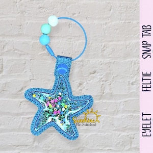 May include: A blue starfish-shaped feltie with glitter and colorful confetti, attached to a blue loop with three beads. The feltie has a stitched outline and the text "Sunshine Be-Stitched".