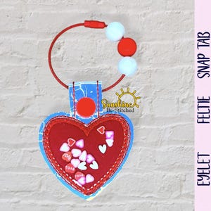 Può includere: Un portachiavi a forma di cuore rosso con un centro trasparente pieno di piccoli coriandoli a forma di cuore. Il portachiavi ha un bordo blu fantasia e un anello con cordino e perline rosse. Il testo "Sunshine Be Stitched" è visibile.